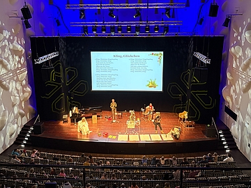 Blick in einen festlich beleuchteten Theatersaal in Potsdam. Auf der Bühne stehen Musikerinnen und Musiker vor einem großen Bildschirm mit dem Liedtext „Kling, Glöckchen“. Im Publikum sitzen viele Kinder und Erwachsene.